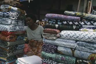 GHANA, Accra, Female vendor framed by piled reams of cloth on stall in Makolo market