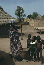 GAMBIA, Agriculture, Woman pounding grain in village with children gathered around her