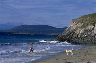 IRELAND, County Galway, Galway, Beach with man walking out of surf towards a dog on the sand