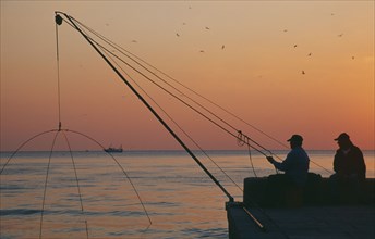 ITALY, Tuscany, Castiglione della Pescaia, Fishermen on harbour wall at sunset with distant fishing