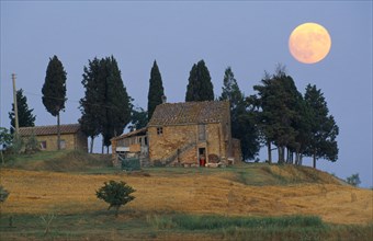 ITALY, Tuscany, Full moon in evening sky over farmhouse and cypress trees near Volterra.