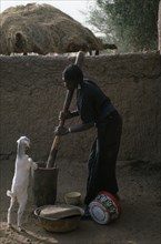 NIGER, Agriculture, Young woman pounding grain with goat kid standing up on hind legs against