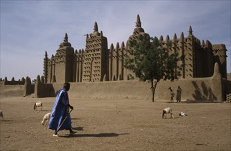 MALI, Sahel, Djenne, Grand Mosque with man walking past and sheep in the foreground.