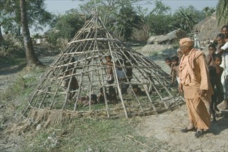 INDIA, West Bengal, Local guru or holy man on village tour inspecting frame of hut waiting to be