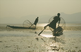 MYANMAR, Shan State, Inle Lake, Silhouettes of two Intha fishermen with Sun’s reflection on water.