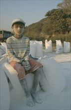 BURMA, Mandalay , "Portrait of a young boy worker at Mandalay Dock sat on bags of cement covered in