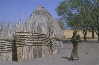 SUDAN, South, Woman and child in Shilluk village walking past thatched hut and enclosure.