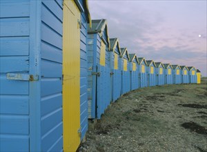 ENGLAND, West Sussex, Littlehampton, Crescent of blue and yellow beach huts in evening light with