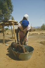 ZIMBABWE, Hygiene, Washing, Woman carrying child on her back washing another in tin bath on farm.
