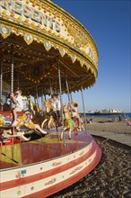 ENGLAND, East Sussex, Brighton, Children riding on a fairground carrousel on the beach with