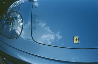 TRANSPORT, Road, Cars, Detail of blue Ferrari emblem and front light with clouds reflected in