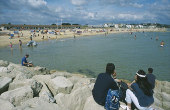 ENGLAND, Dorset, Sandbanks, People sat on rocks looking across water and busy sandy beachfront