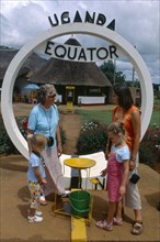 UGANDA, Equator, European family watching water go down plughole beside sign.