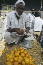 INDIA, Markets, "Man behind display of oranges on stall, crouching on ground holding money."