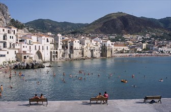 ITALY, Sicily, Palermo, Cefalu. View from stone walls with people sat on benches towards sunbathers