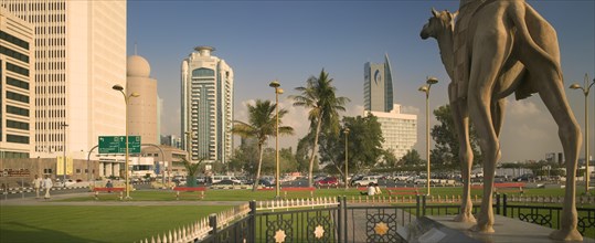 UAE, Dubai, "Camel statue in Al-Ittihad Square, people sat on grass. Palm trees and car park in