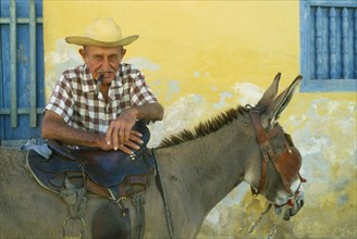 CUBA, Trinidad, Portrait of elderly Cuban man smoking a cigar and leaning on saddle of blinkered