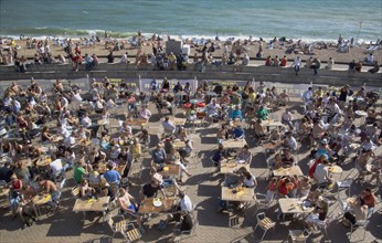 ENGLAND, East Sussex, Brighton, People sat at tables drinking outside the Beach seafront bar.