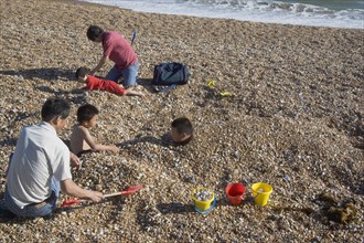 ENGLAND, East Sussex, Brighton, Family on the beach with the children being covered in pebbles.