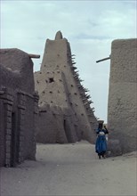 MALI, Tomboctou, Street scene with mud built  Mosque and woman carrying a bowl on her head.