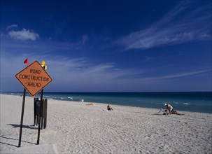 USA, Florida, Fort Lauderdale Beach, Road Construction Ahead sign on sandy beach with sunbathers