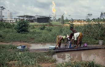NIGERIA, Environment, Women washing clothes in foreground with oil refinery behind.