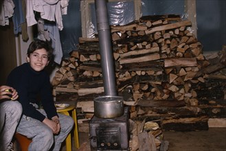 ARMENIA, Yerevan, Boy sitting beside wood burning stove in house with large stack of wood behind