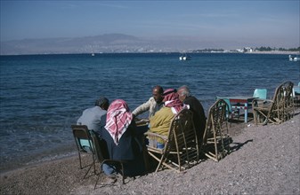 JORDAN, Aqaba, Group of men playing backgammon on shore of beach with Eliat and Israel in distance