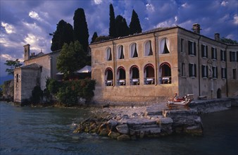 ITALY, Lombardy, Lake Garda , Punta San Vigilio.  Villa on lake shore with couple on sun loungers