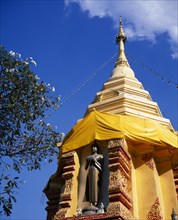 THAILAND, North, Chiang Mai, Standing Buddha statue below gold chedi wrapped in golden orange silk