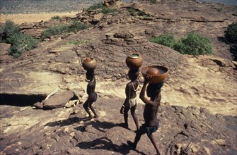 20088820 MALI Pays Dogon People Dogon boys carrying water pots on their heads over rocky terrain.