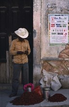 MEXICO, Oaxaca, Man standing beside crumbling plaster wall with pile of red chillies for sale at