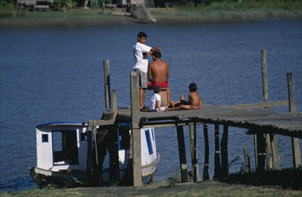 20088598 BRAZIL Para Aflua Amazon Estuary.  Man having a haircut while waiting for the ferry on wooden jetty watched by two children.