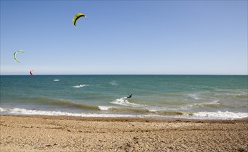 ENGLAND, West Sussex, Lancing, Kite Surfers on sea next to shingle beach in the summer with blue