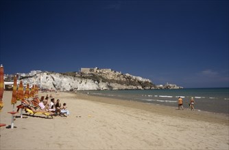 ITALY, Puglia, Foggia, Vieste.  Line of sunbathers on stretch of sandy beach overlooked by castle