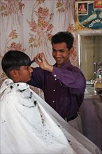 INDIA, Rajasthan, Jodhpur, Young Indian barber in his barbershop cutting the hair of a boy.