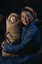 MONGOLIA, People, Young mother in fleece-lined silk tunic holding baby wrapped in traditional