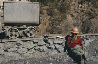 BOLIVIA, Chukiuta, Miner wearing orange hard hat sitting next to mining rail track chewing leaves