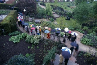 ENGLAND, East Sussex, Glyndebourne, Opera attendees walking through the gardens holding umbrellas