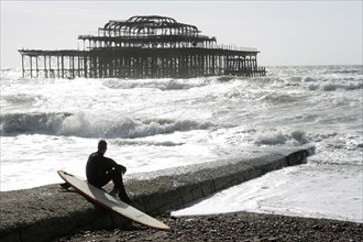 ENGLAND, East Sussex, Brighton, "Seafront, man sat with surf board on goyne in front of the burnt