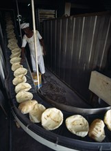 Qatar, Doha City, Baker in bakery standing beside conveyor belt carrying round Arab breads coming