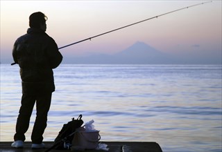 Japan, Honshu, Chiba, Tateyama, man fishing in Tokyo Bay, Mount Fuji visible across the water.