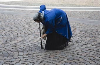 Budapest, Pest County, Hungary. Elderly woman bent over and supported with cane begging in square