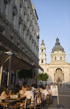 Budapest, Pest County, Hungary. Cafe with Summer tourists seated at outside tables with Saint