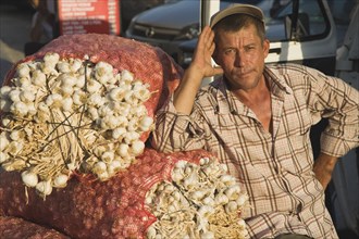 Kusadasi, Aydin Province, Turkey. Stallholder at weekly market leaning on sacks of garlic in the