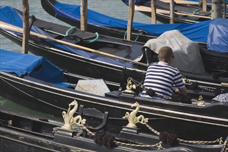 Venice, Veneto, Italy. Gondolier wearing striped shirt using mobile phone sitting in one of a line