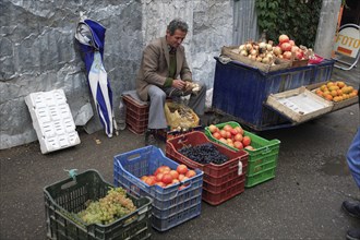 Albania, Tirane, Tirana, Fruit and vegetable street vendor seated on crate to bunch garlic behind