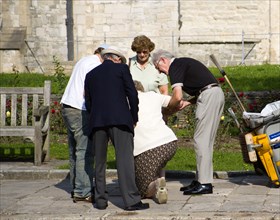 People helping an elderly lady to stand up after she has fallen on the pavement sidewalk outside