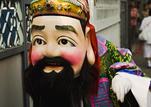 Man in Chinese costume with papier-mache head and beard carried in parade celebrating local temple.