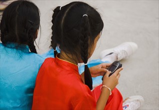 Thai girl using mobile telephone seated with friend in costume of dance troupe.